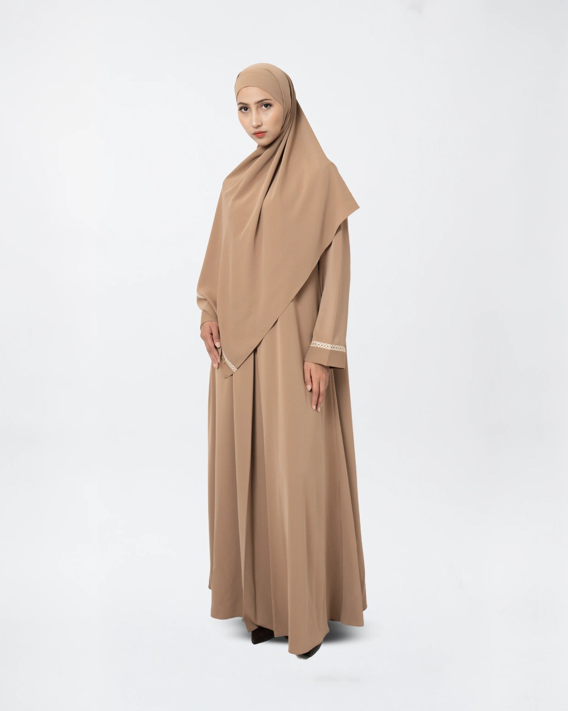 A woman modeling a modest outfit consisting of a long beige abaya dress with a draped hijab, standing against a plain white studio background.